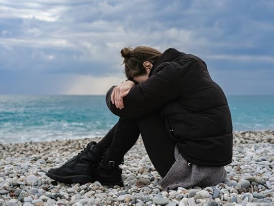 A woman sitting sadly on a pebbly beach. You can find positivity when it feels like it all sucks 