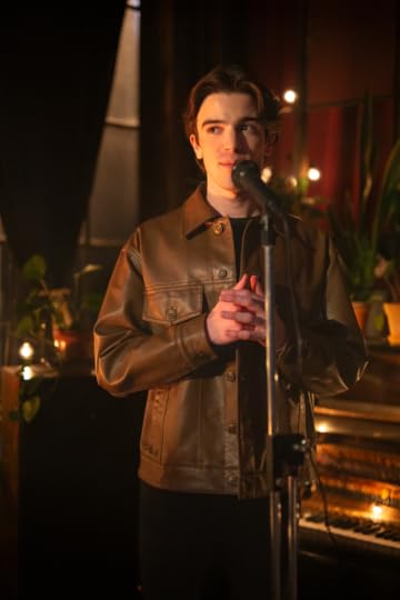 A man wearing a brown leather jacket stands at a microphone in a dimly lit bar
