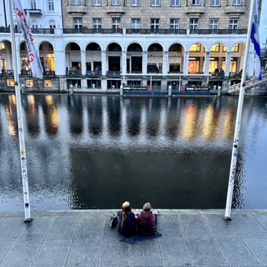 Ein Paar sitzt auf dem gepflasterten Ufer der Kleinen Alster und sieht auf die Alterarkaden