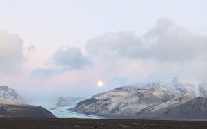 A bright blue glacial lagoon surrounded by gray mountains, with a full moon rising in the background