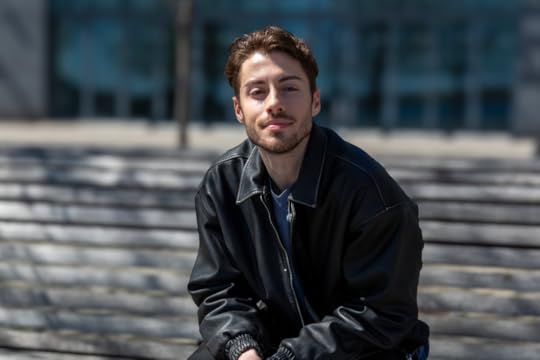 A man in a black leather jacket sits outside on a bench with his hands folded in his lap