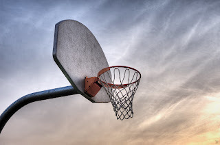 Image of an outdoor basketball hoop with white backboard, orange rim, and nylon net. Sky is at sunset