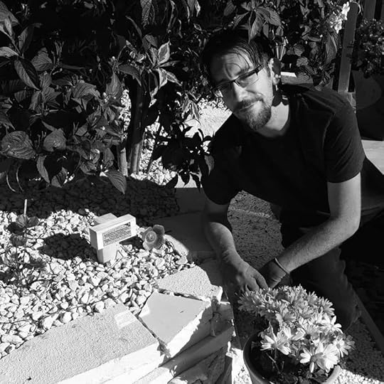black and white author photo of the author in a garden. He is a white man with glasses and short dark hair, a trimmed beard and moustache. He is wearing a short sleeved black t-shirt.