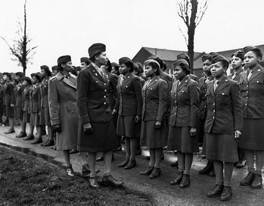 A group of uniformed women in military attire stand in formation on a road. Leafless trees and buildings line the background. A woman, who appears to be inspecting them, wears a long coat and cap. The scene is formal and emphasizes discipline and uniformity, indicative of a military context.
