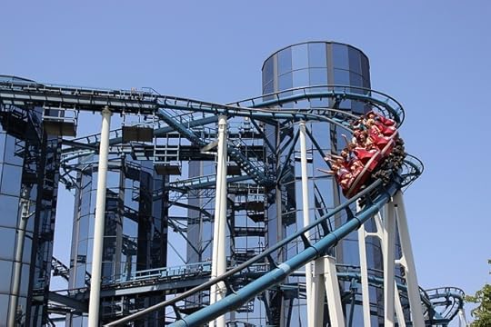 Industrial looking tall, round roller coaster with a car full of people rolling around and downward, their hands up in the air.