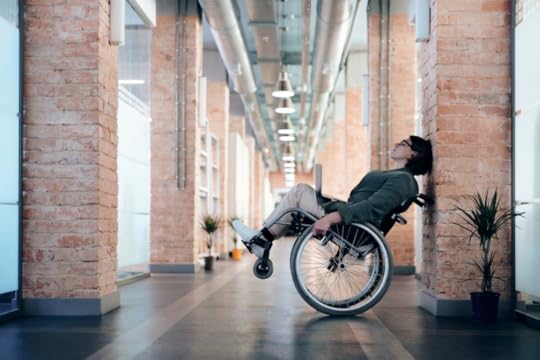 Image: a woman with a computer on her lap leans her wheelchair back against a brick column in a brightly lit modern office building.