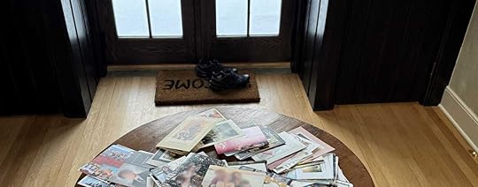 A round wooden table in the center of a room holds a large pile of holiday cards, some overlapping and scattered. The background features a set of dark wooden doors with glass panes, showing a snowy outdoor scene with a wooden deck and chairs. A doormat with the word 