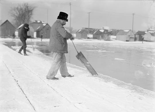 Black and white image of a man with a long-handled ice saw cutting ice on the ege of a narrow lake or frozen river.