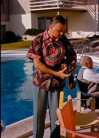 A man taking a photograph standing next to a resort swimming pool. He is wearing gray slacks and a brightly colored print shirt. 