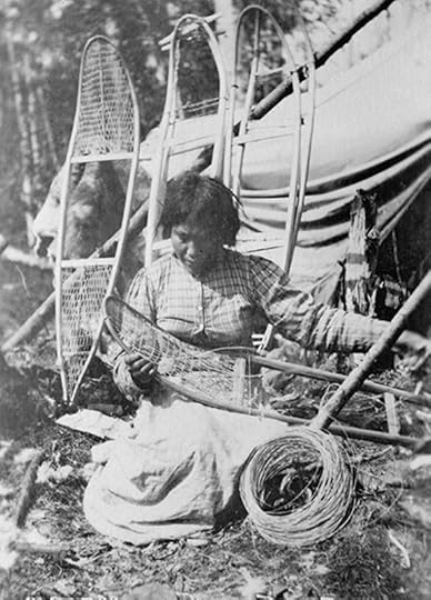A Native American woman, seated, weaving the netting in a pari of snowshoes. She is sitting outside with tress and possibly a tent just behind her.