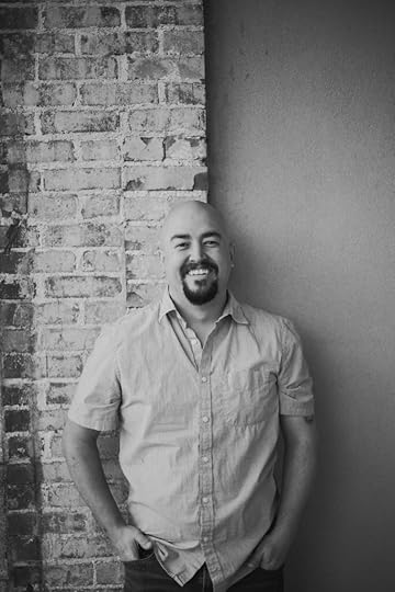 author photo of Chris Coles standing against a brick wall background. Ze is a bald white person with a moustache and trimmed beard cropped around zis chin, and is wearing a casual combination of a light coloured shirt and dark trousers. Zis photo is in black and white. Ze is smiling.