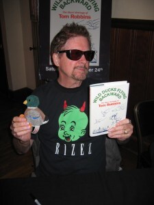 Tom Rbbins holding a stuffed dick and a copy of his short stories titled Wild Ducks Flying Backwards