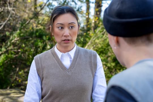 A woman wearing a sweater vest and button down shirt sits opposite a person wearing a beanie
