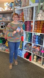 Photo of Jo at The Wool Cellar holding The Natter of Knitters in front of shelves full of wool