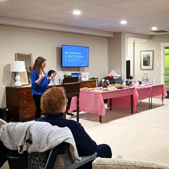 I stand at a wooden podium, speaking to an audience in a cozy room. A screen behind me displays the presentation title: 