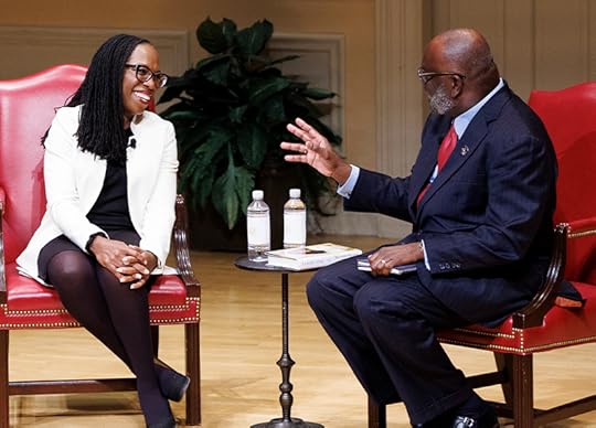 A well dressed man and a woman, seated in elegant red leather chairs, share a laugh on stage. 