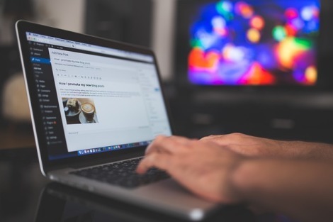 Hands typing on a laptop with a multi-colored out of focus tv screen off to one side