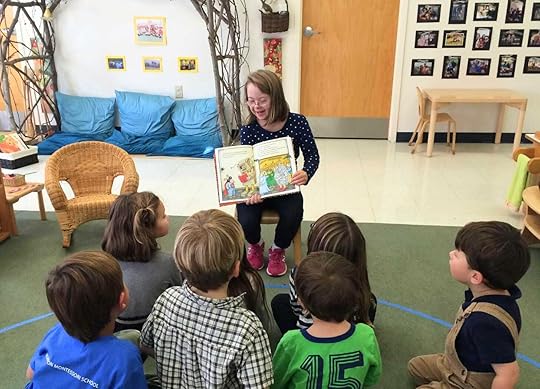 Penny in 2015 sitting in a small chair in a classroom and reading a book to students.