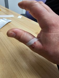 PIcture of a thumb held over a wooden effect laminate desk with a cut that has a steri-strip across it.