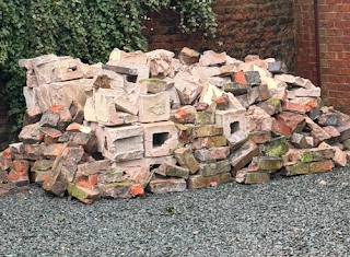 A pile of bricks - house bricks and breeze blocks - are piled up outside a brown garage door. There's a wall of green ivy behind, and the drive way is covered in grey slate.