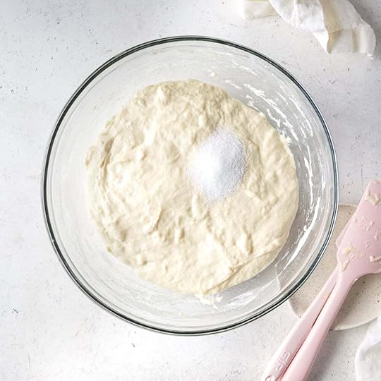 Puffy focaccia dough with salt on top in a large bowl with pink spatula. 