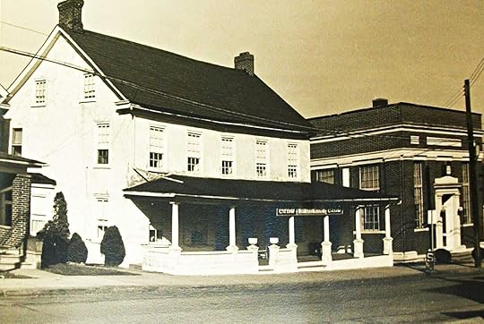 Facing empty street, white 2.5 story building with front sloped roof and porch next to institutional building with white doorway.