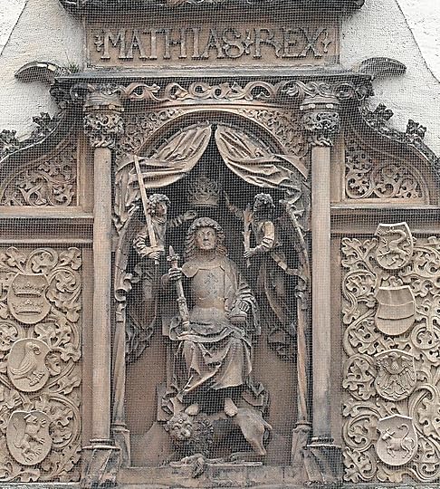 Impressive relief sculpture of Matthias Corvinus on a wall in Bautzen Ortenburg. The kin wears armor and has angels holding a crown above his head, one angel with a sword the other with an olive branch. The king holds a scepter and has his feet on a lion. Around him are carved ornate columns and vines, and above the inscription