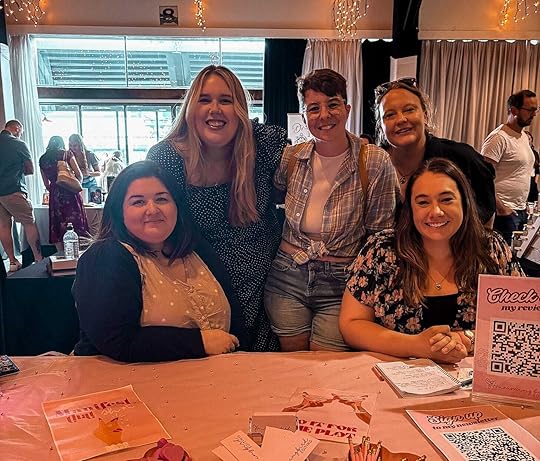 Five people in a photo standing and sitting behind a vendor stall. The stall belongs to Hummingbird Editing. The people behind the stall from left to right are: Laura Ibic (Hummingbird Editing), MG Hunt, Laura Catherine, Krystal from Aussie Indie Reads, and SK May.