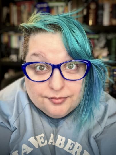 Author & artist Raven Oak with green eyes, blue/teal hair (with a side shave), blue shirt, and blue glasses. They are sitting in front of their office library.