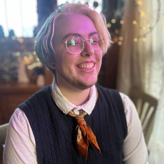 A blond person with short hair and glasses, some light facial hair, smiling widely in a cosy room lit by fairy lights and natural light. They are wearing an orange silk scarf knotted like a short tie, a white shirt and a sleeveless knitted navy sweater over the top.