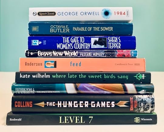 Pictured is a stack of books including George Orwell's 1984, Octavia Butler's Parable of the Sower, Sheri Tepper's The Gate to Women's Country, Aldous Huxley's Brave New World, M.T. Anderson's Feed, Kate Wilhelm's Where Late the Sweet Birds Sang, Frederik Pohl and C.M. Kornbluth's The Space Merchants, Suzanne Collins's The Hunger Games, and Mordecai Roshwald's Level 7.