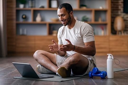 Fitness coach/personal trainer in a gym sits in front of his computer on a yoga mat next to dumbbell and water bottle