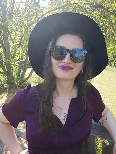 white woman in black hat and large sunglasses, purple shirt, outside in the sunshine with green trees behind her.
