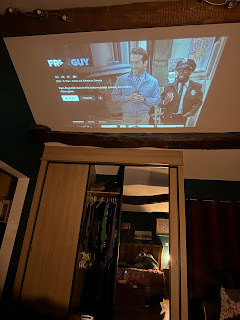 A dark bedroom. There are wardrobes, with a mirror and then above that, a sloping ceiling. There is a projection of the Disney + information for the film Free Guy.