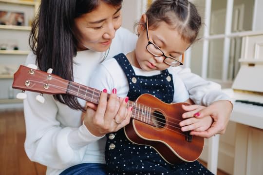 Mother guiding her young daughter with glasses as they learn to play the ukulele together at home