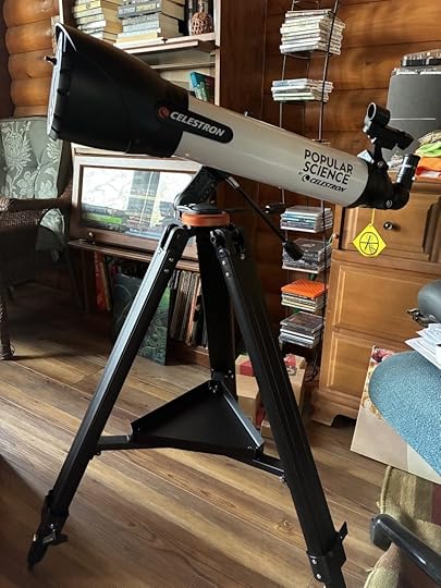 A telescope mounted on a tripod set up in the center of a somewhat-cluttered living room.