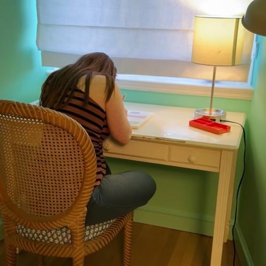  Penny sits at a wooden table in a softly lit room. She is writing in a notebook. A small table lamp with a white shade provides light. A red pencil case sits on the desk near the lamp.