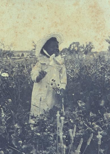 A grainy, sepia-toned photo of a young girl in a field, holding a few flowers. She is wearing a white dress and a large bonnet with a strap tied in an elaborate bow under her chin.