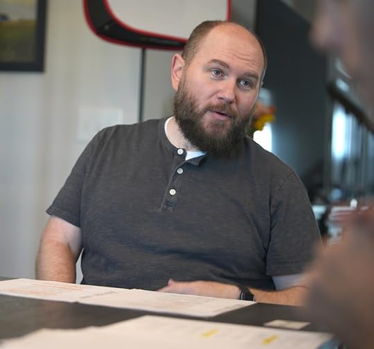 stocky white man with brown beard seated at a table, leaning sideways as if midway through a line of dialogue, with a script in front of him.