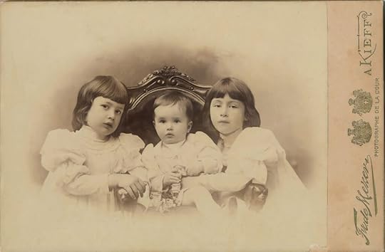Old sepia photo of three small children, two girls and a baby boy, in Nineteenth Century clothes posed sitting in a chair.