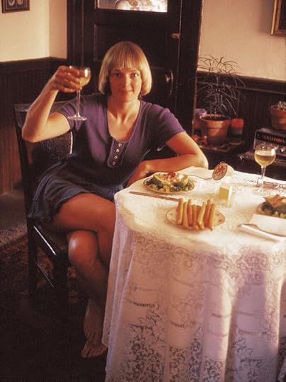 Young blond woman wearing a purple dress and holding a glass of wine. She is sitting at a small table with a long lace tablecloth and plates of food.