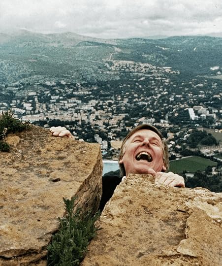 Man pretending to scream while hanging by his fingers to rocks over a landscape of green hills and houses. Meant to be a funny image.
