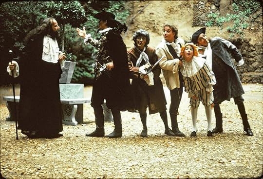 A group of five actors in Elizabethan costume lined up and listening in a Shakespearean comedy let outdoors with autumn leaves on the ground.