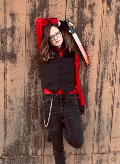 NR Wolfman's author photo, full length against a fence in black jeans, red shirt and black waistcoat. Light-skinned woman with glasses and brown hair, holding a fake-bloodied prop knife in a casual pose, arm behind her head.