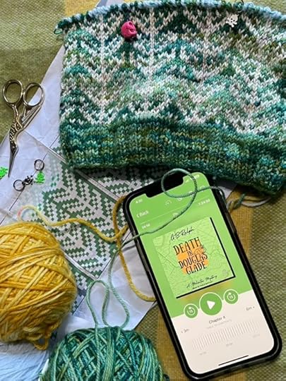 Knitting in progress alongside balls of green and orange wool, and a phone showing the Death in Douglas Glade audiobook by G. B. Ralph.