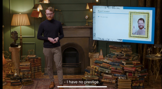 Ryan Creamer from Pornhub standing in a room presenting a slideshow. he is a red headed and bearded male, dressed in black round glasses, black long sleeved turtleneck and khaki slacks with brown dress shoes. His slide is a photo of him shirtless in a presumed shower with a blank white screen to his left for a drop-down list. in the photo (in the presentation) he is giving a thumbs up. The slide is titled 'Ryan Creamer'. Ryan presents this slide while stating "I have no prestige"