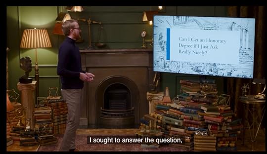 Ryan Creamer from Pornhub, a red headed bearded man presenting a slideshow. He is wearing glasses, a turtleneck black shirt, khaki slacks and brown dress shoes. The first slide in the presentation is Titled 'Can I get an Honorary Degree if I Just Ask Really Nicely' 