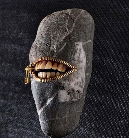 A sculpture by Hirotoshi Ito, of what appears to be a river-smoothed stone with a brass zipper on it. The zipper is open, revealing smiling human teeth.