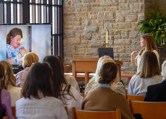 I’m giving a presentation in a warmly lit room with stone walls and wooden furniture. I stand at a podium, gesturing as I speak to an audience of women seated in rows of chairs. A large screen beside me displays an image of me in a hospital bed holding my newborn Penny, with hospital identification bracelets visible on my wrist. 
