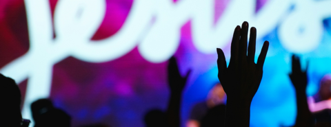 Silhouettes of hands raised in front of a neon Jesus sign during a Praise & Worship event